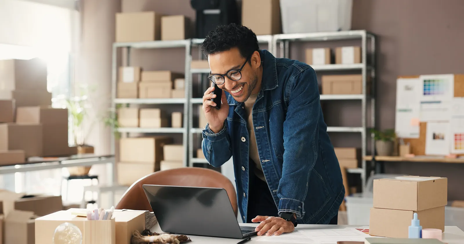 Small business owner taking a call at his laptop in a shipping workspace